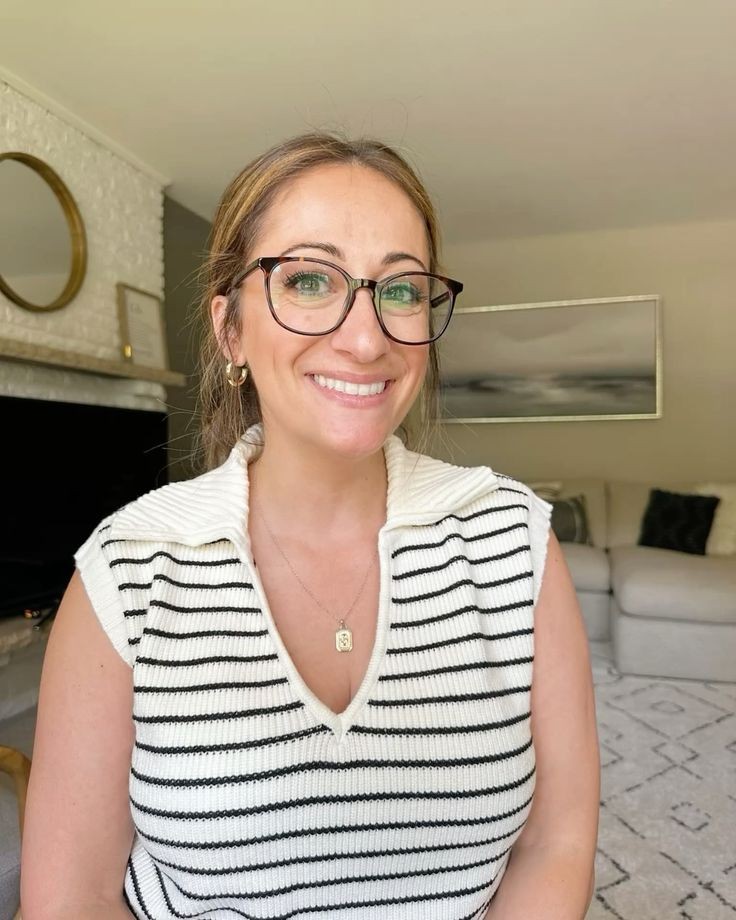 Smiling woman with glasses, wearing a striped sweater, seated in a living room with a sofa and abstract art on the wall.