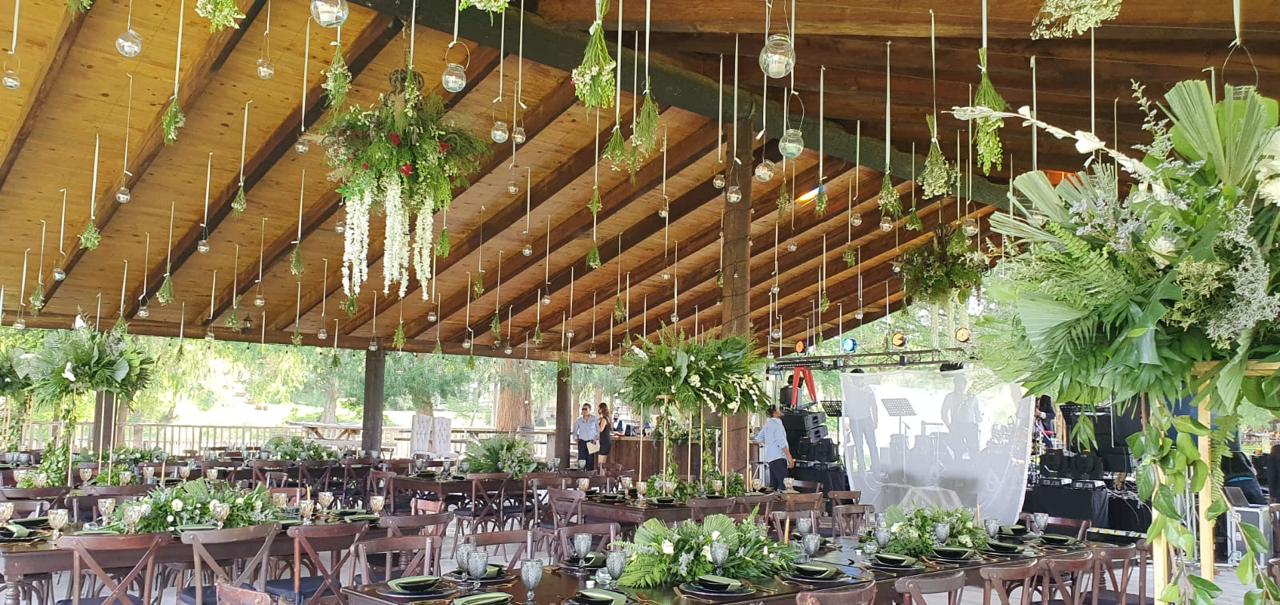 Rustic wedding reception with wooden tables, green floral arrangements, and hanging greenery under a wooden roof.