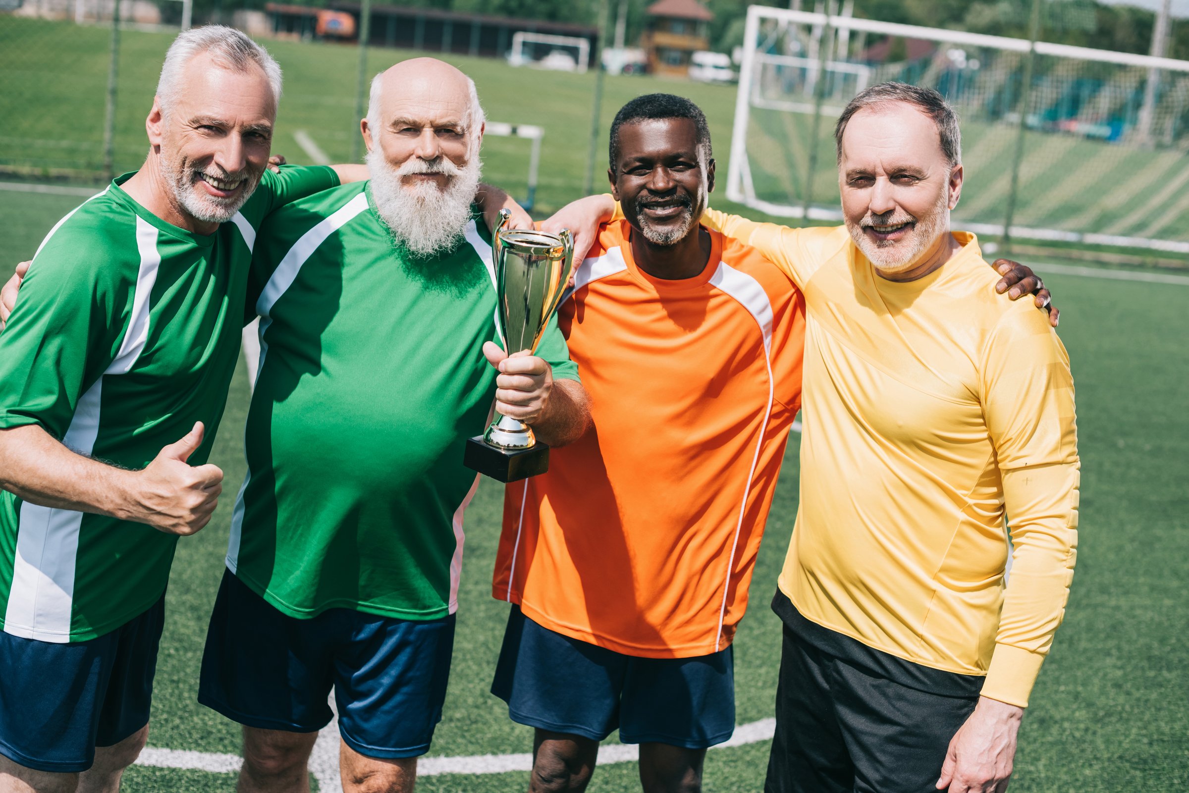 multiethnic smiling old sportsmen with champions cup standing on football field
