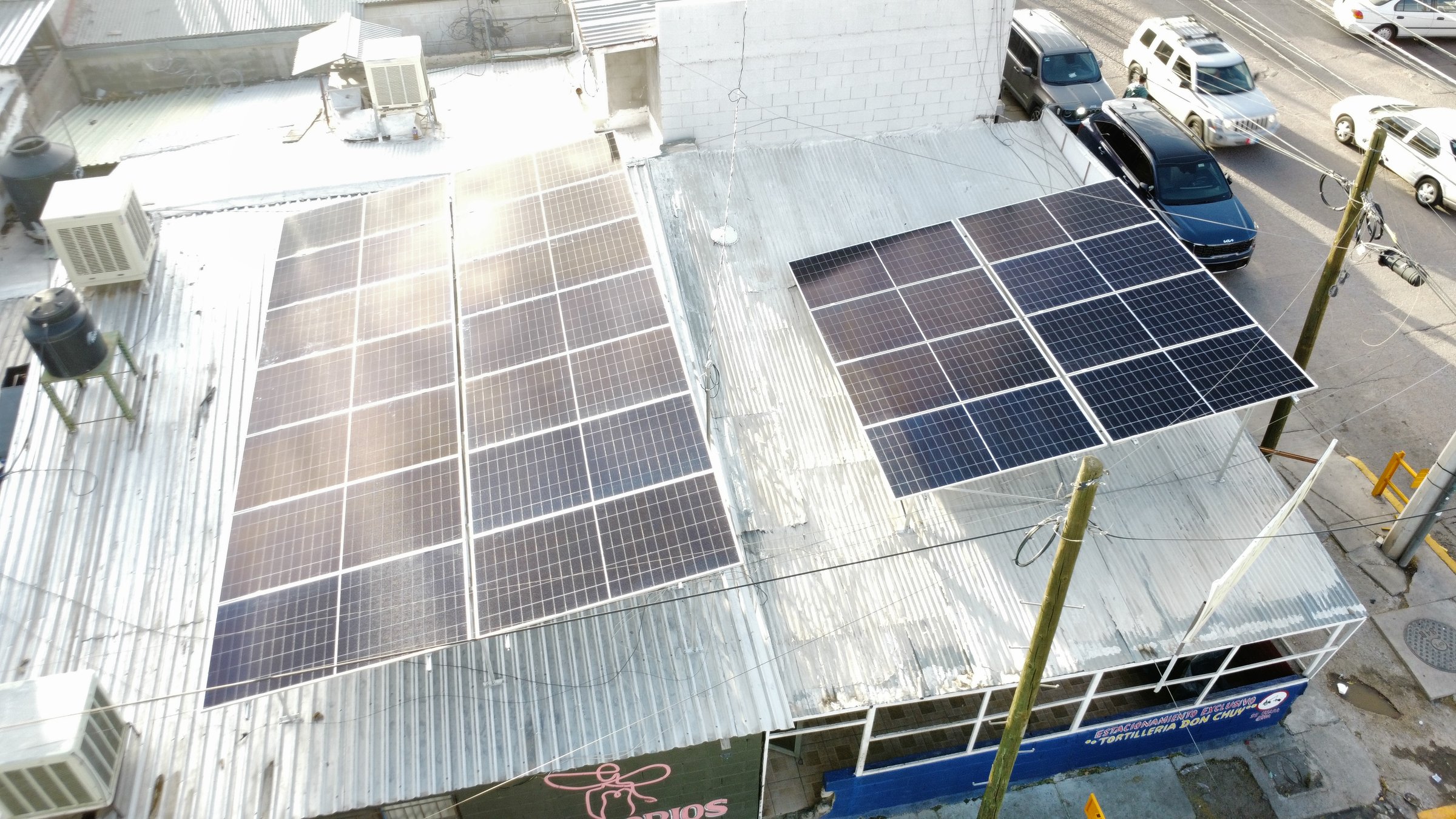 Aerial view of a building with solar panels on the roof, surrounded by cars and utility poles on a sunny day.