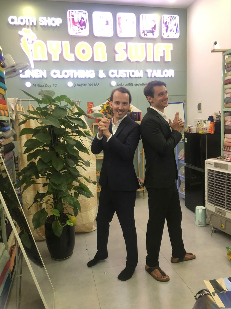 Two men in suits pose playfully as secret agents in a tailor shop named "Taylor Swift Linen Clothing & Custom Tailor."