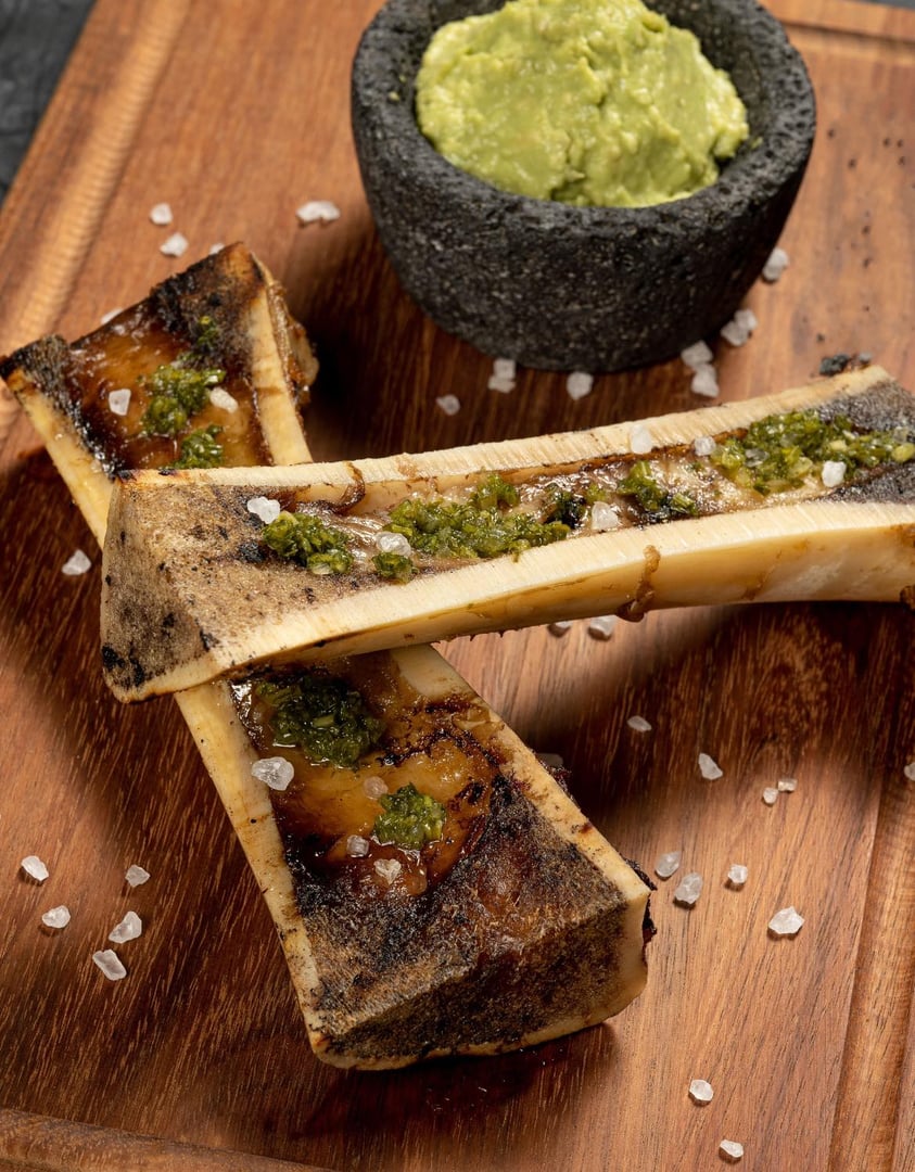 Grilled bone marrow with herbs and coarse salt, served on a wooden board with avocado dip in a stone bowl.