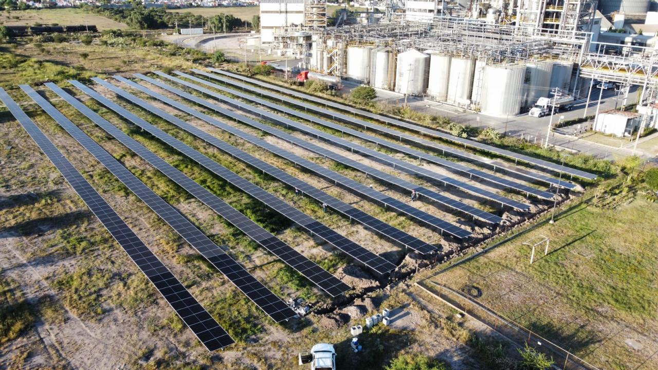 Solar panels installed in rows on a grassy field near an industrial complex with tanks and structures in the background.
