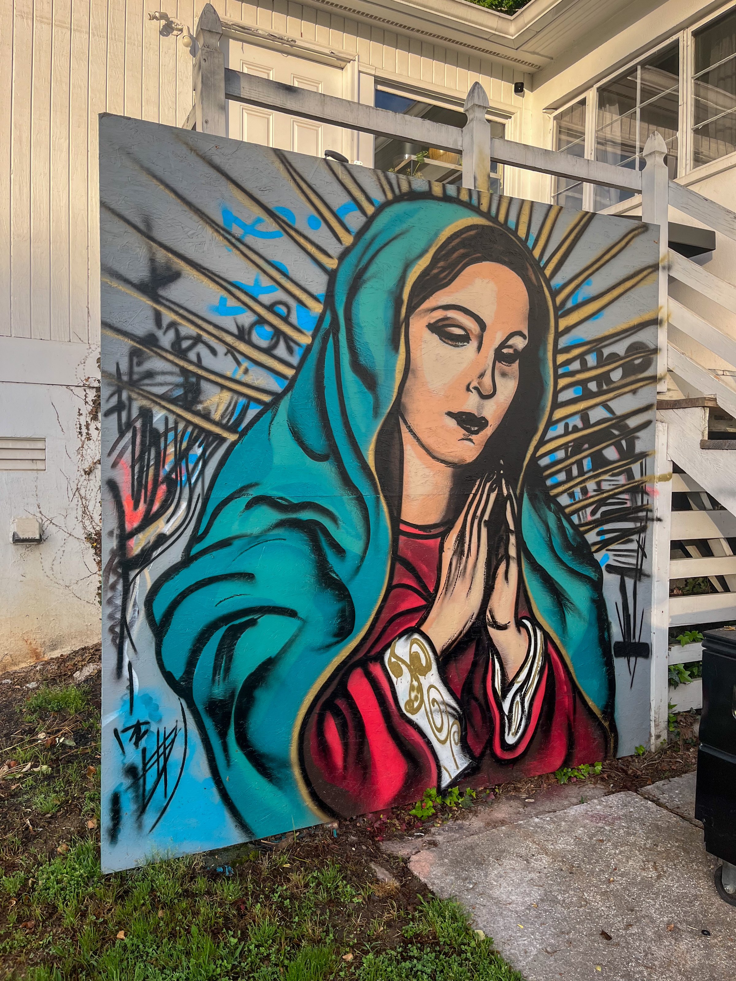 Colorful mural of a praying woman in a blue robe, with rays emanating from her head, painted on a wooden panel.