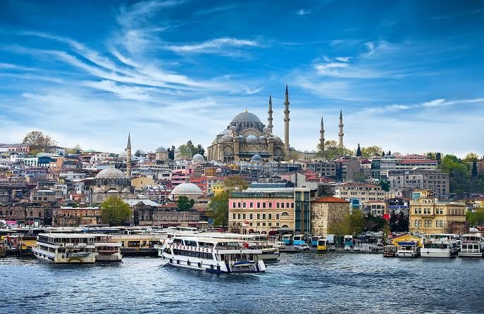 View of Istanbul's waterfront with boats, multi-story buildings, and a prominent mosque under a blue sky.