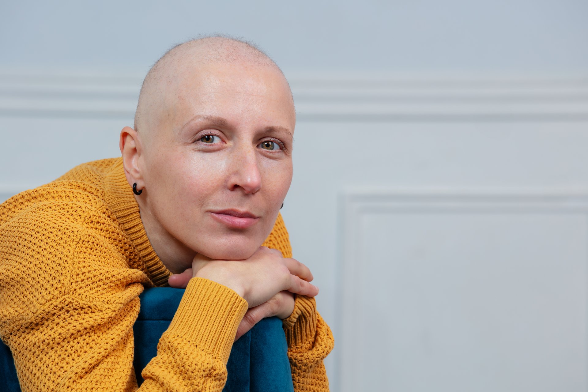 A bald woman cancer survivor with serene expression sitting in a chair, her hand resting on its arm, a white wall in the background