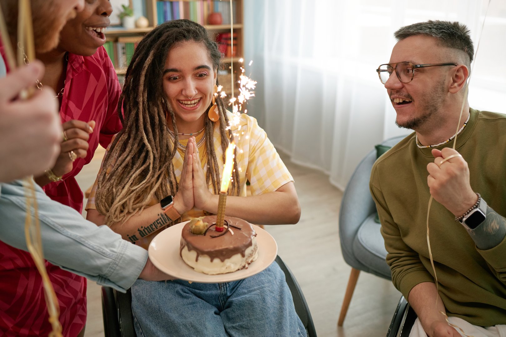 Multiethnic group of young adults with disability celebrating birthday, smiling and clapping while sitting in wheelchairs, woman with dreadlocks holding cake with sparkler candle