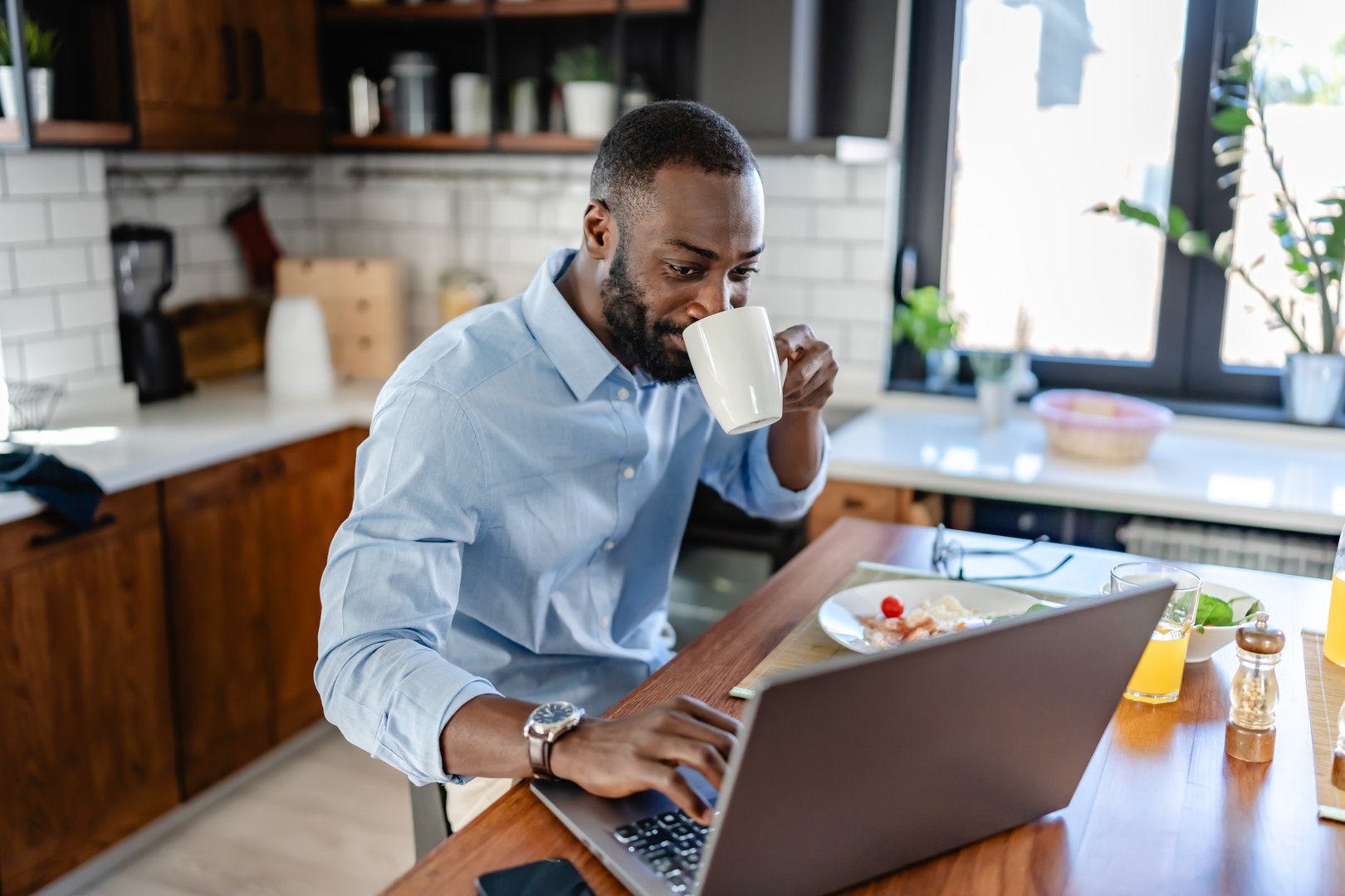 African-American man in a modern kitchen, drinking coffee and working on a laptop during breakfast. Bright and productive morning scene.