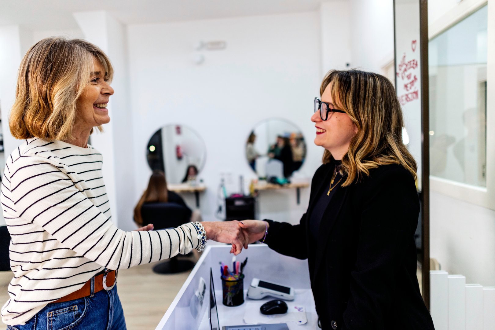 Two smiling women are shaking hands at the reception desk of a modern hair salon