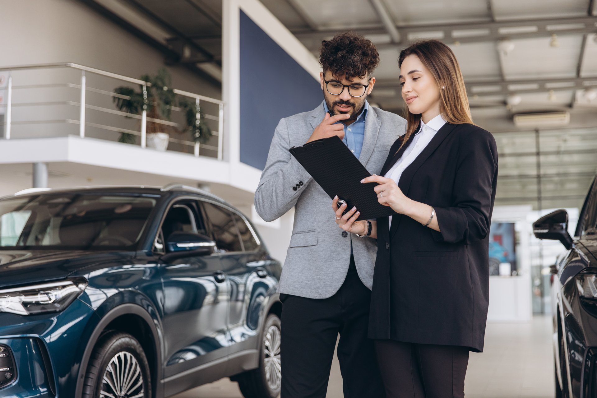 Salesperson explaining car features to a thoughtful client holding a clipboard, engaging in a detailed discussion inside a modern dealership