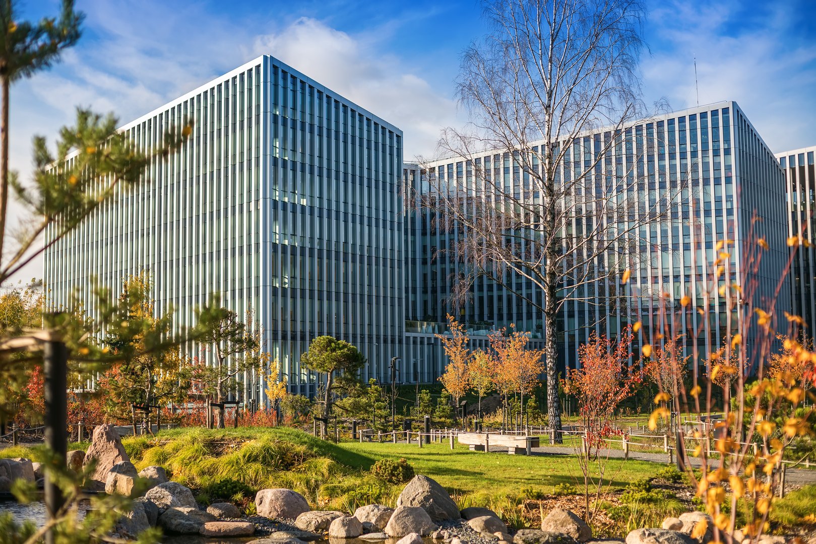 The modern glass facade mirrors the sky, standing beside a landscaped park with autumn trees, lush grass, and a small pond, combining urban design with natural beauty