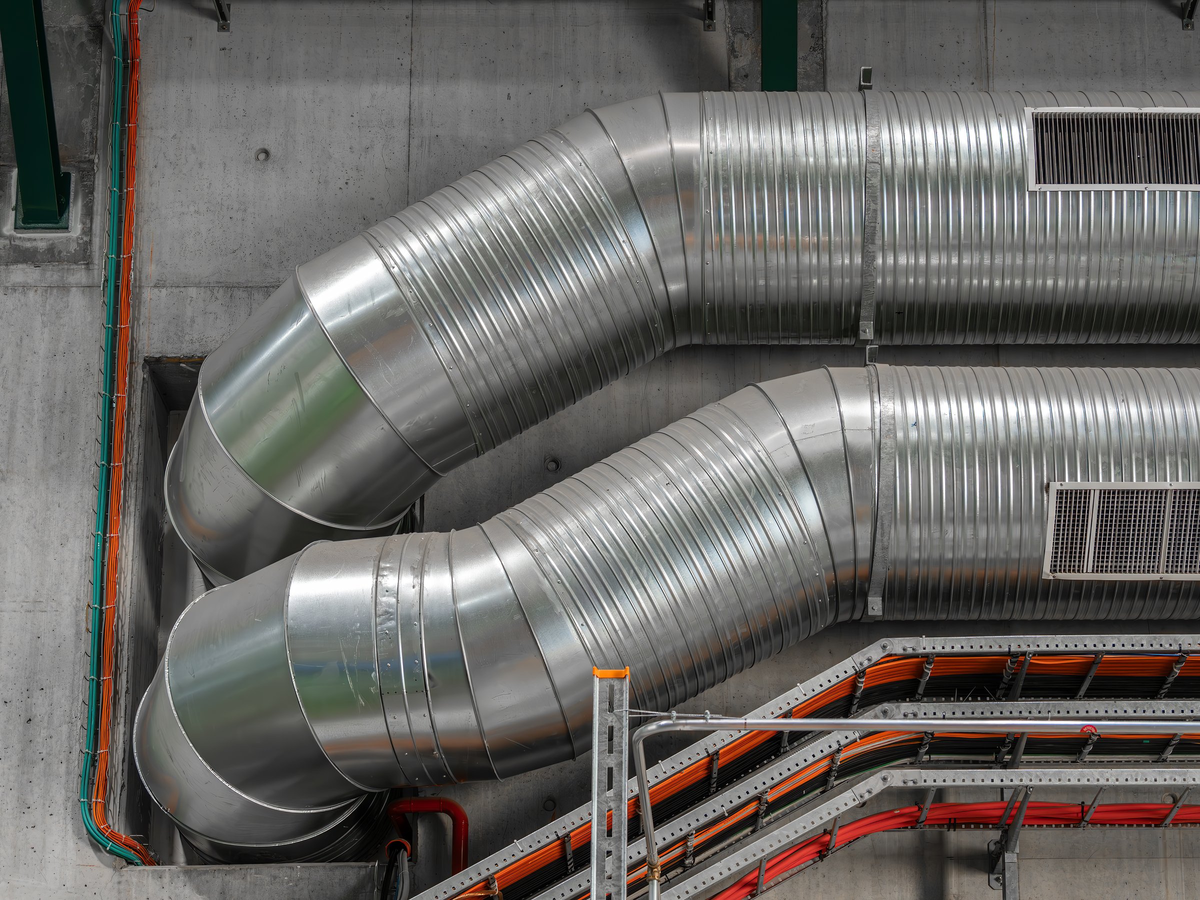 Close-up of large metallic ducts and pipes in an industrial setting, showcasing their shiny surface and intricate design against a concrete wall.