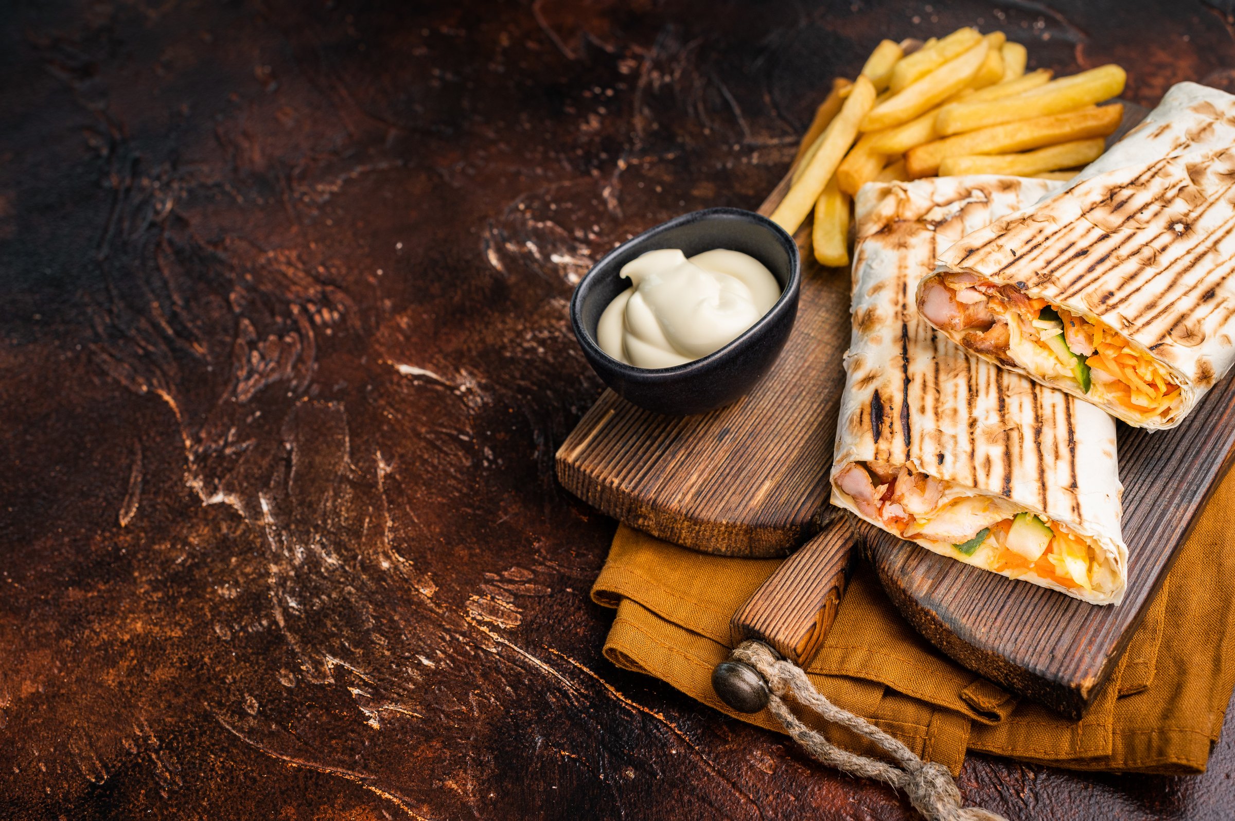 Shawarma chicken roll with fresh vegetables, cream sauce and french fries on wooden board. brown background. top view.
