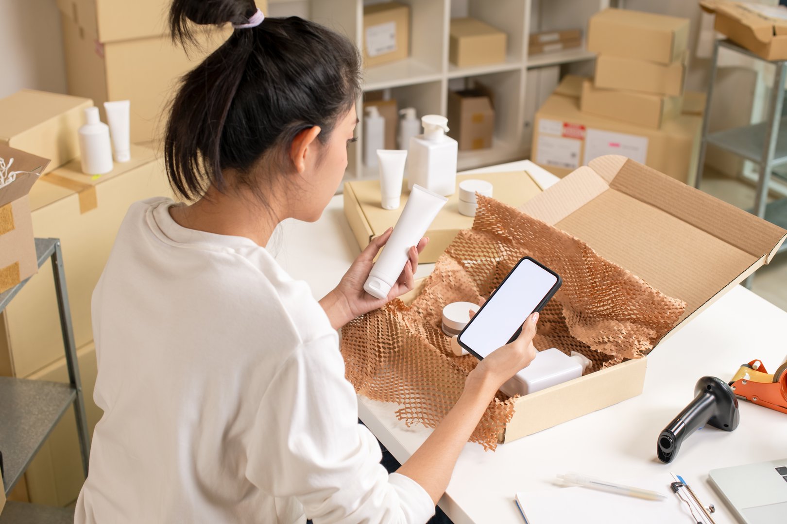 Asian e-commerce seller inspecting and sorting products for customers, using mock-up app, in a warehouse setting for an online beauty shop.