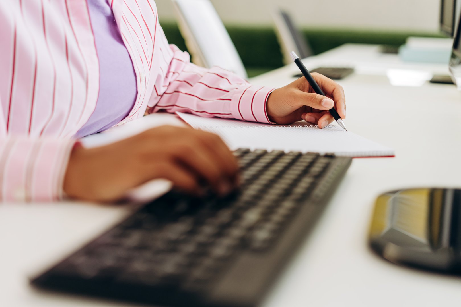 Close-up of hands working on computer keyboard and writing notes in spiral notebook, showcasing multitasking and productivity in modern office environment