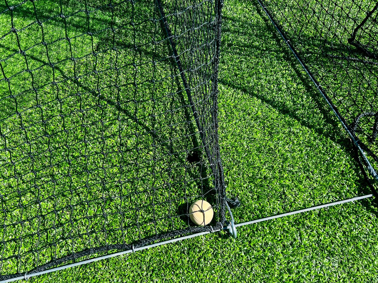 Low angle view of an enclosed batting cage with black netting on green artificial turf. A baseball is tucked underneath of the the batting cage nets.