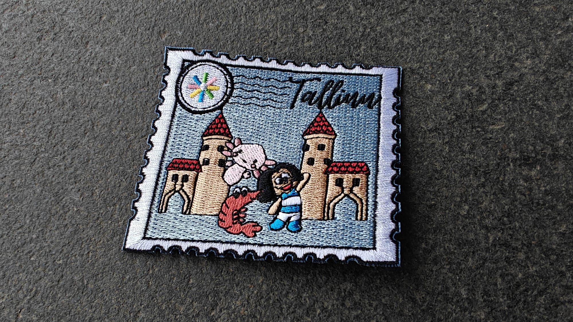 Embroidered patch resembling a stamp with cartoon castles, a cow, and a person, labeled Tallinn in black script.