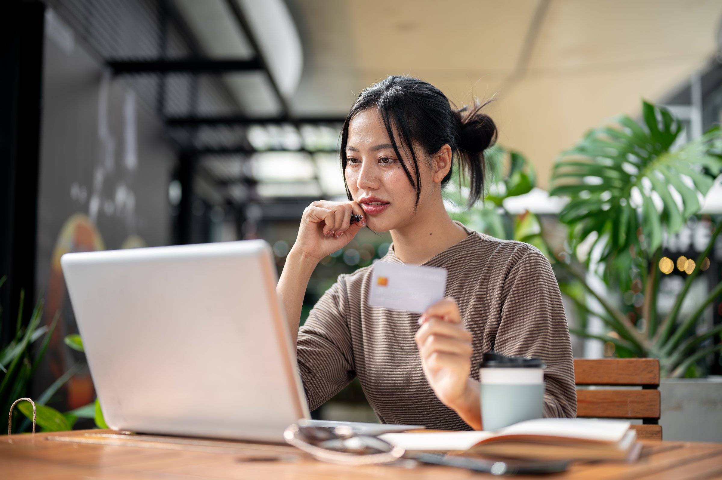 A charming, happy Asian woman is paying bills online or shopping online through her laptop while sitting at an outdoor table of a cafe on a bright day. cashless payment, online banking, financial