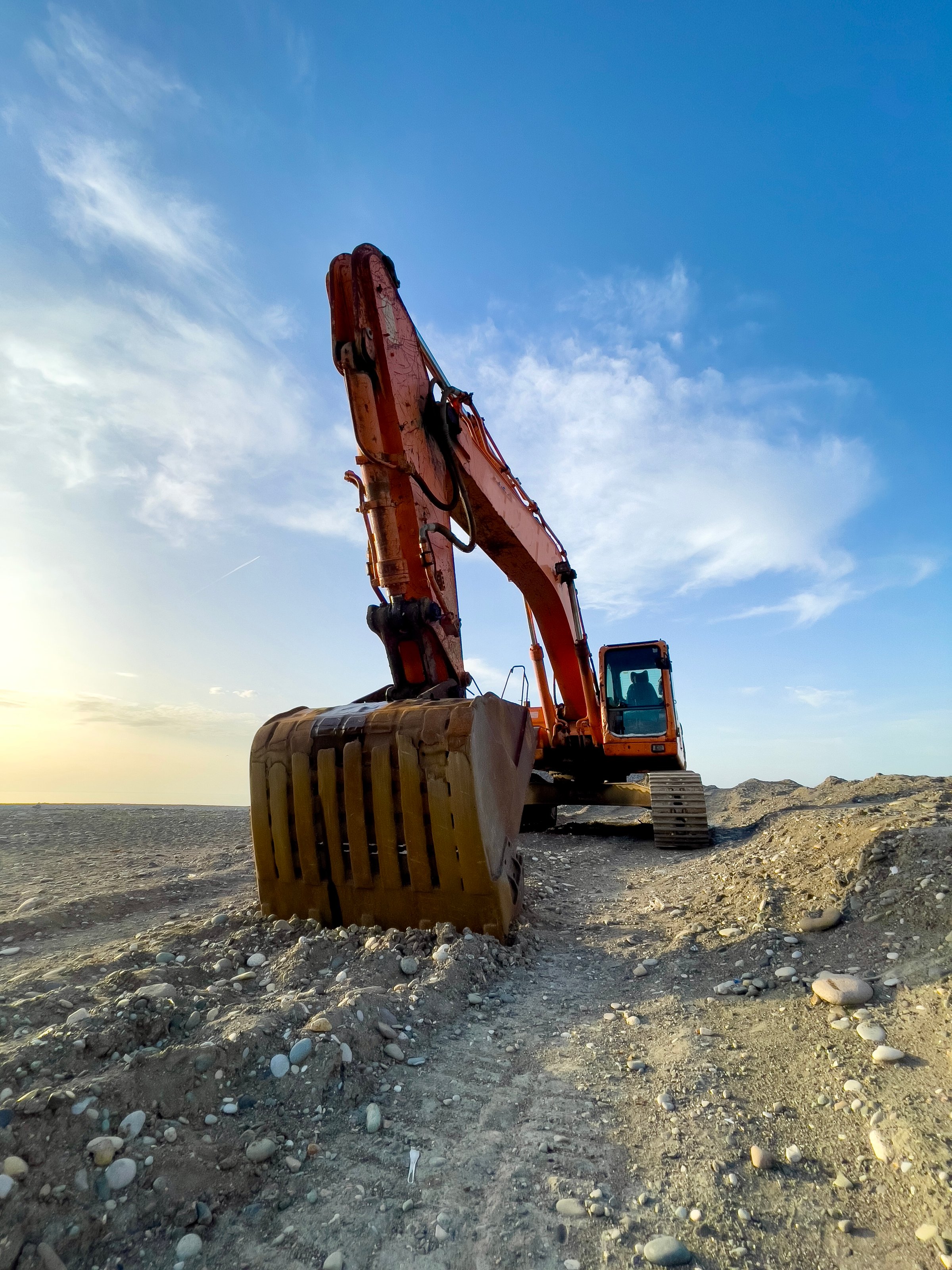 This dynamic image showcases a large orange excavator equipped with a robust bucket attachment, actively engaged in earth-moving activities. Positioned prominently on rocky terrain, the excavator's detailed hydraulic arm and hoses highlight the machinery's engineering complexity and functionality. The bucket, worn from use, adds an authentic touch to the industrial scene. In the background, a vivid blue sky with soft white clouds enhances the visual appeal, creating a sense of openness and productivity. Ideal for themes related to construction, heavy equipment, or engineering projects.