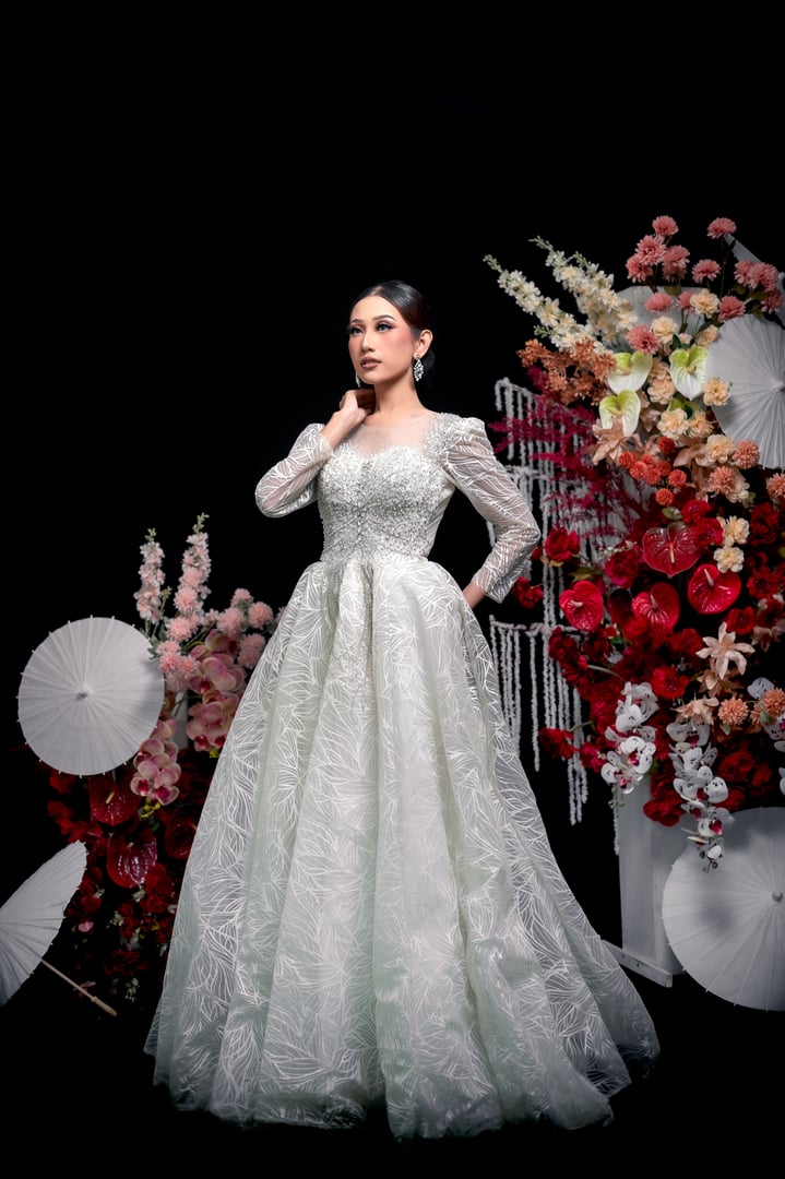 Elegant Indonesian bride in a white embellished gown with pearls and lace details, standing gracefully beside floral arrangements, showcasing flawless bridal makeup and timeless charm.