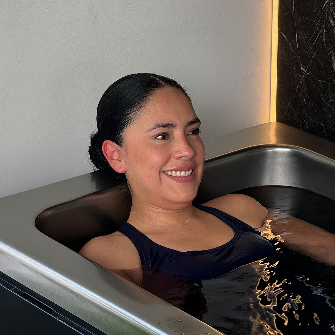 A woman with dark hair in a bun relaxes in a bathtub filled with dark water, smiling softly.