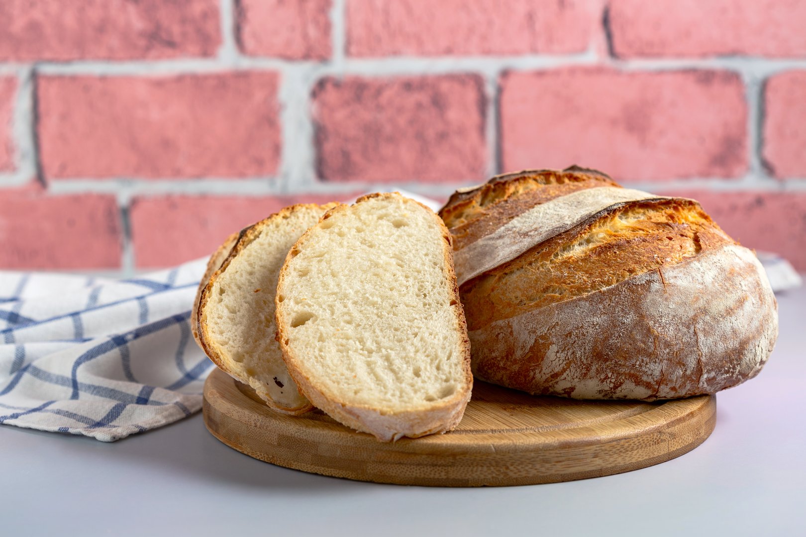 Sliced sourdough wheat bread and a round loaf on a wooden board, with a kitchen towel in the background,