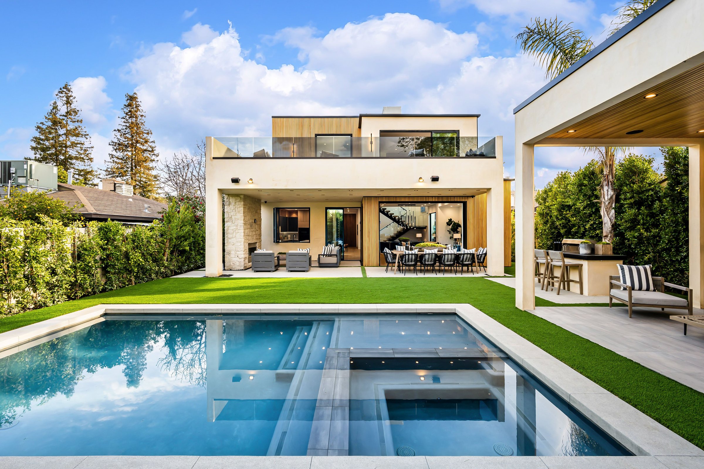Modern luxury house with a swimming pool, outdoor seating area, and lush greenery under a bright blue sky.