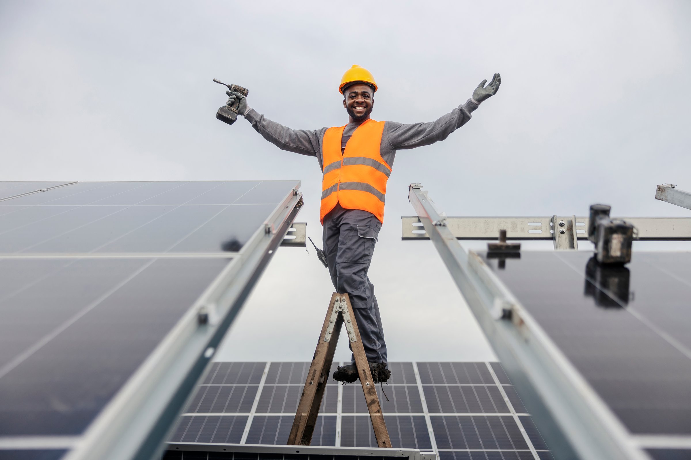 Worker in orange vest and hard hat stands on a ladder, arms spread wide, amidst solar panels, holding a drill.