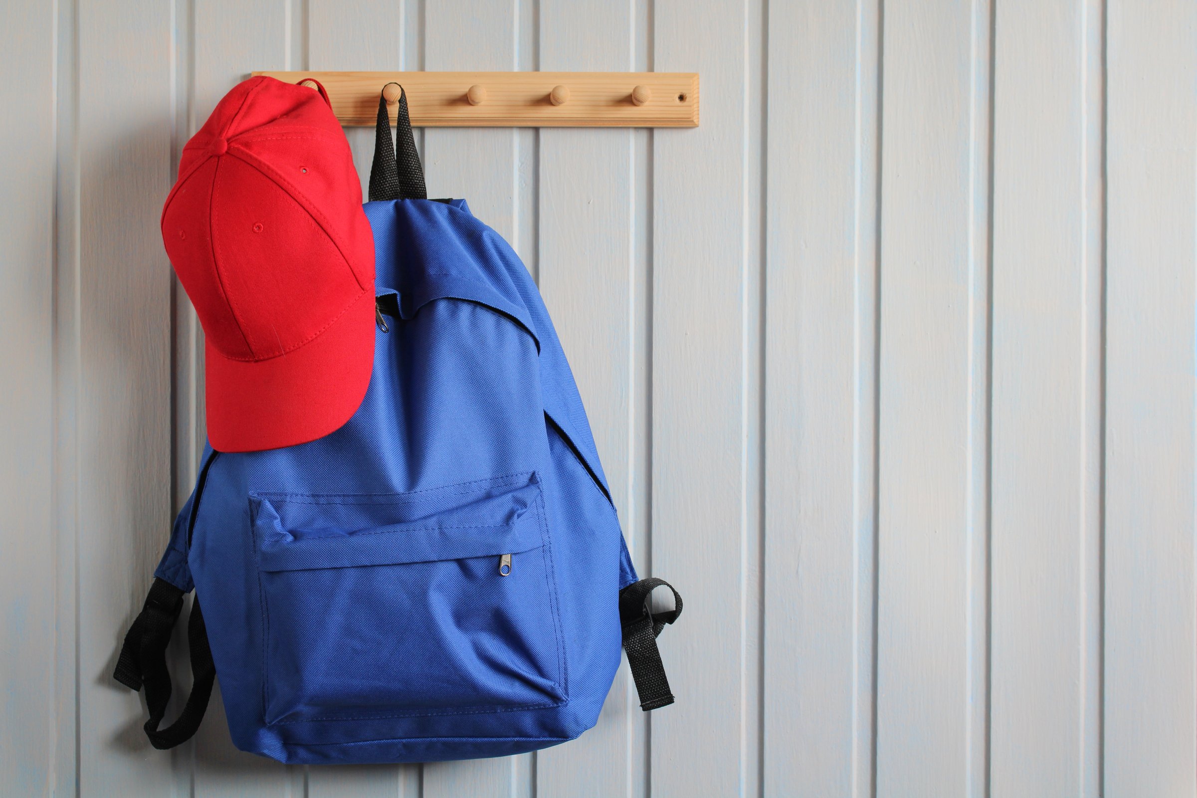 A blue backpack and a red baseball cap hang on a wooden hanger on a white board wall. copy space.