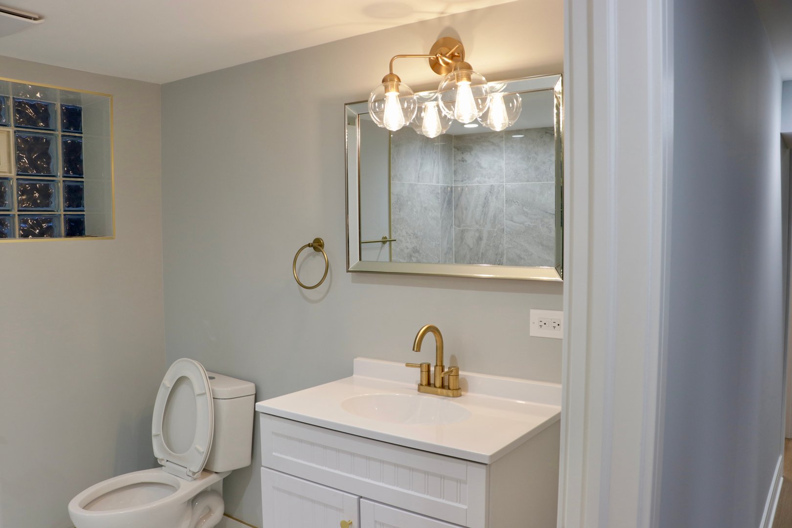 Modern bathroom with a white vanity, brass fixtures, a large mirror, toilet, and glass block window.