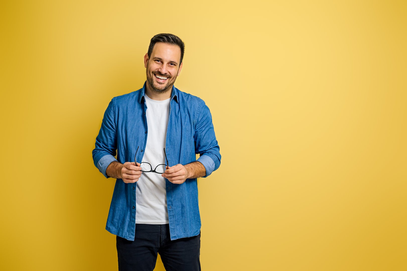 Portrait of smiling handsome businessman holding eyeglasses and standing over yellow background