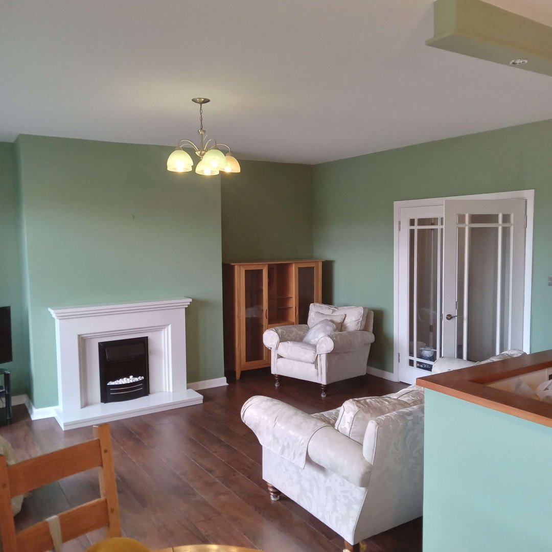 Living room with green walls, a white fireplace, armchairs, and wooden floors. A glass cabinet and chandelier are present.