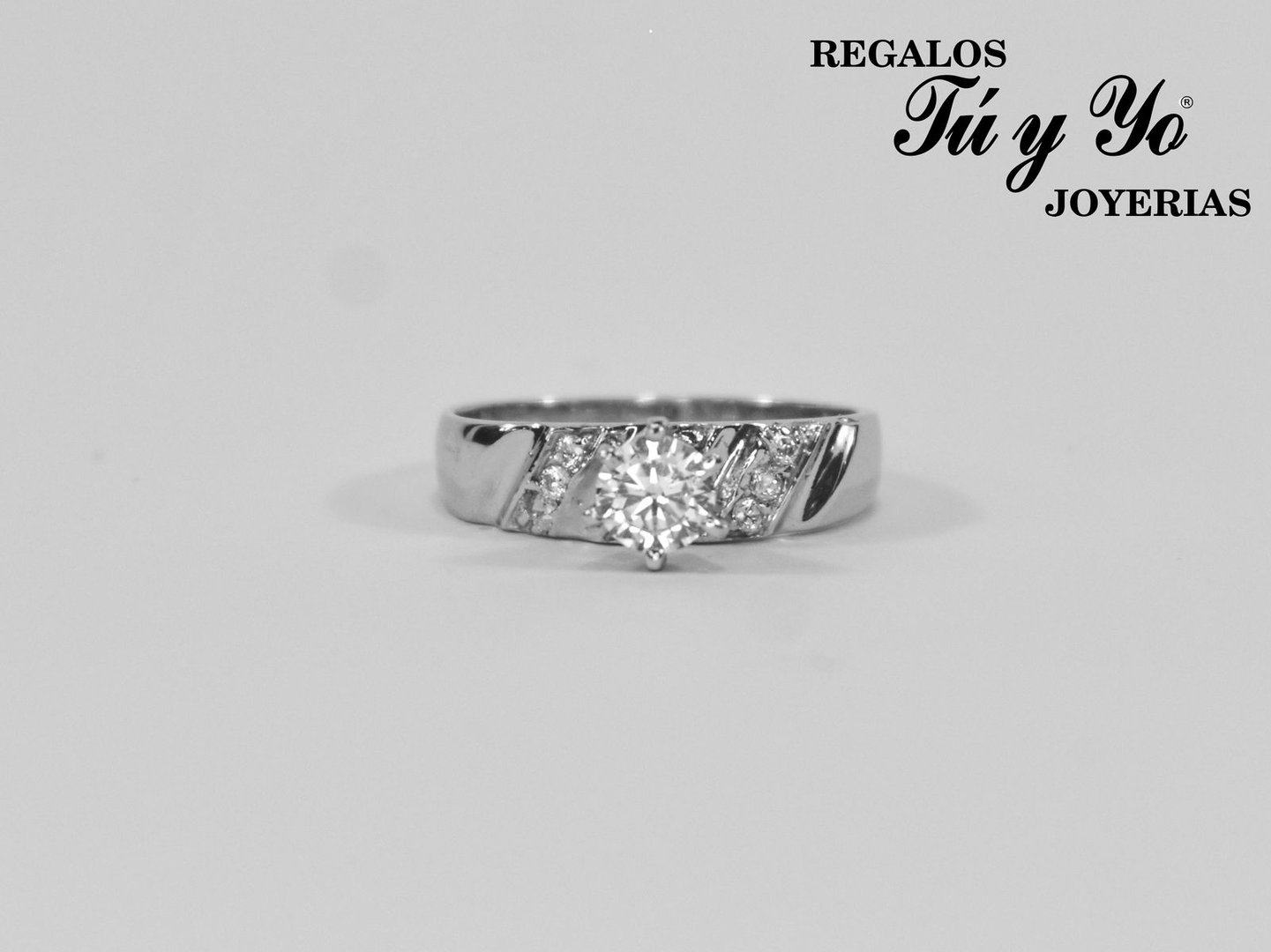 A silver ring with a central diamond and small diamonds on the band, featuring jewelry store branding in the top corner.