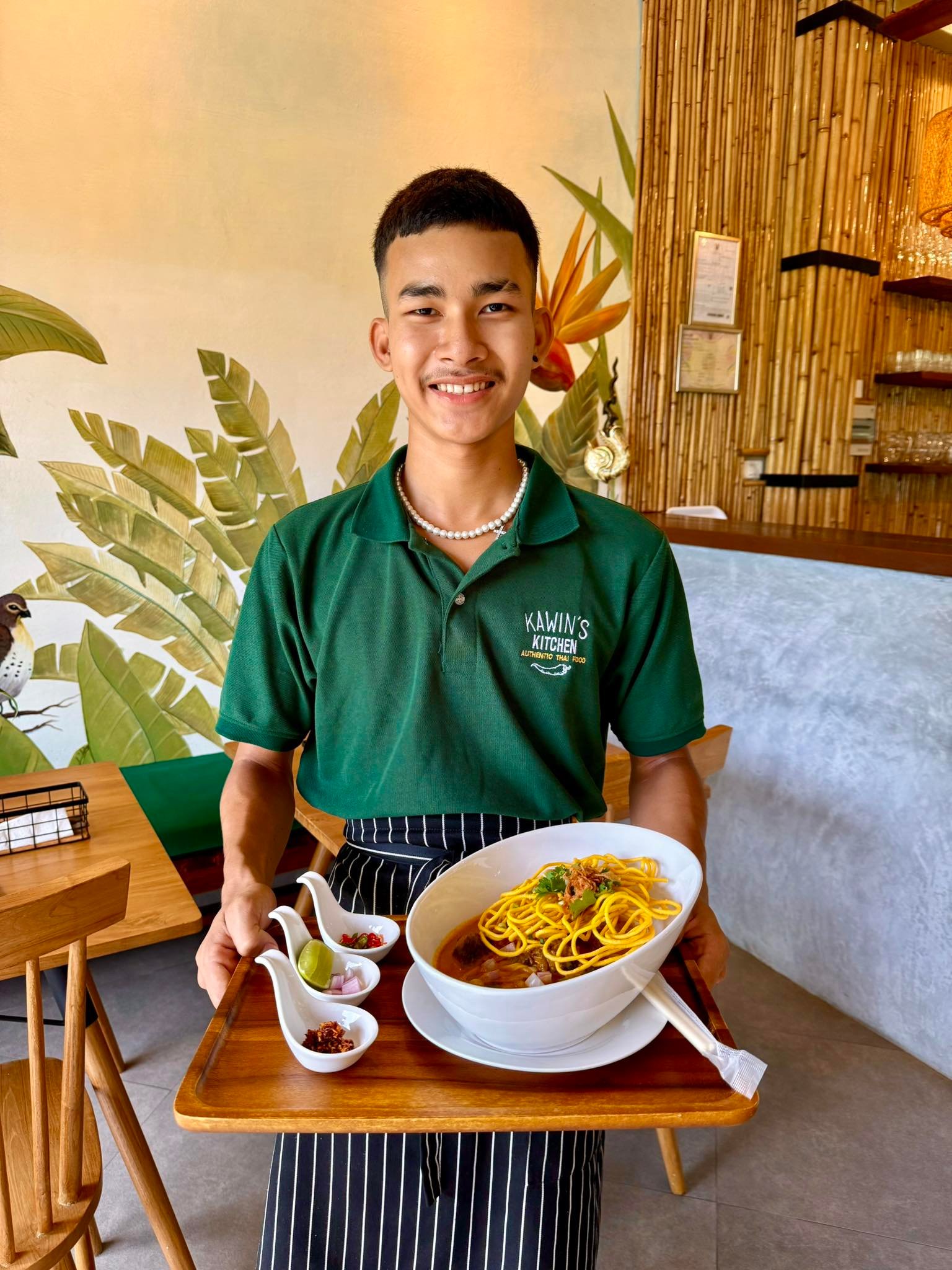 Smiling waiter in green shirt serves a bowl of noodles with condiments on a tray in a cozy restaurant.