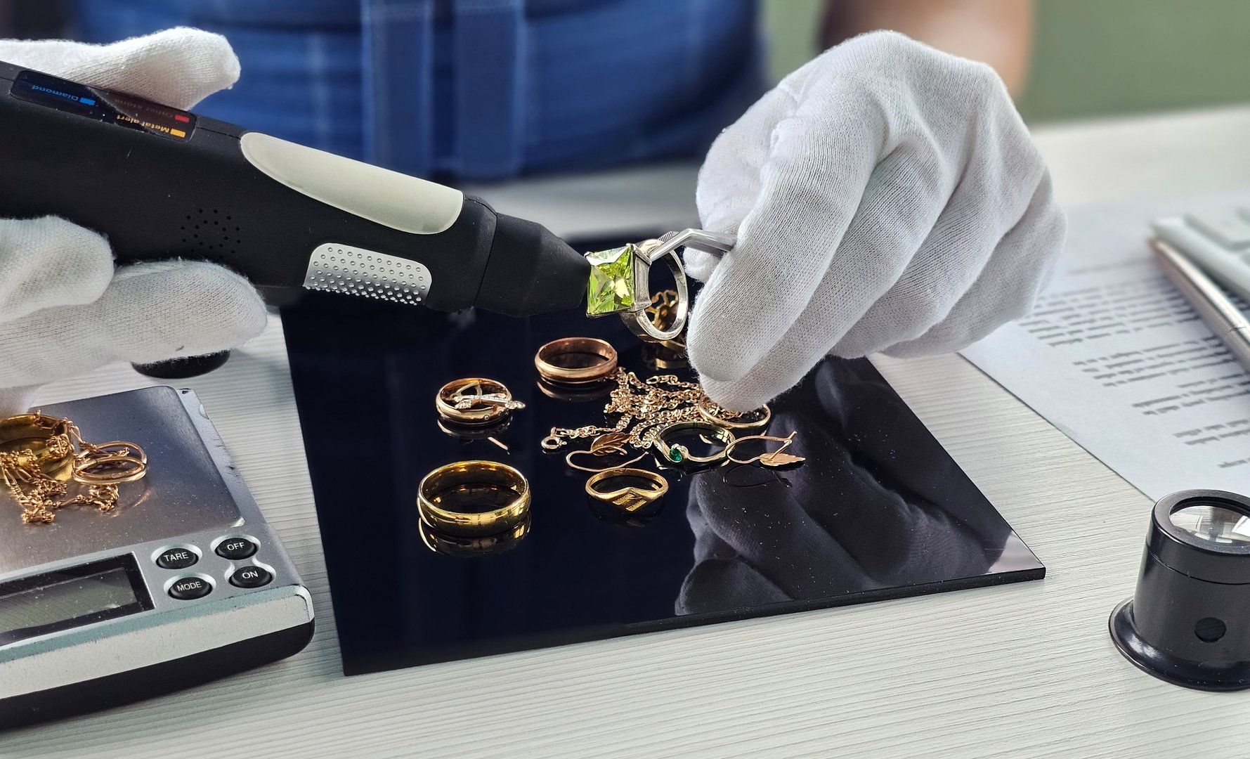 Expert gemologist examines various golden rings and jewelry pieces on a black surface, using a specialized tool for appraisal in a well-lit workshop