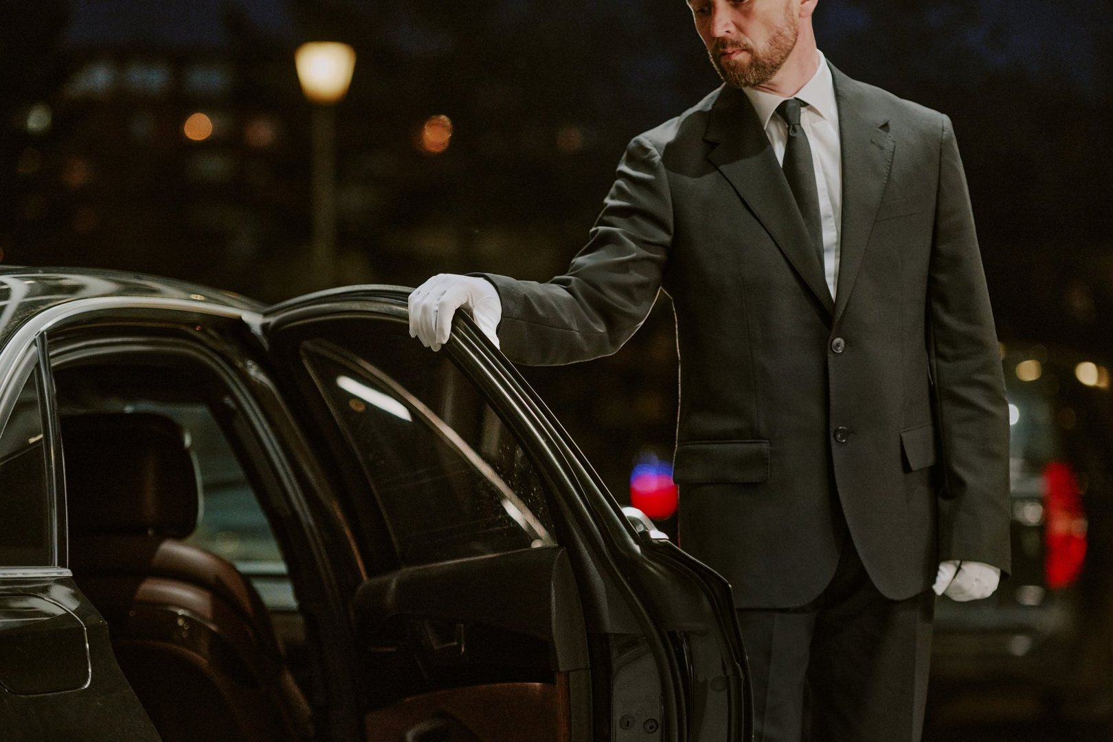 Caucasian middle aged man wearing suit and gloves opening car door outdoors at night, standing beside luxury vehicle, performing professional chauffeur service