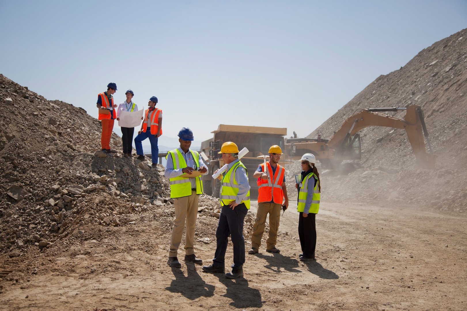 Construction workers in safety gear discuss plans at a quarry site with machinery and piles of dirt in the background.