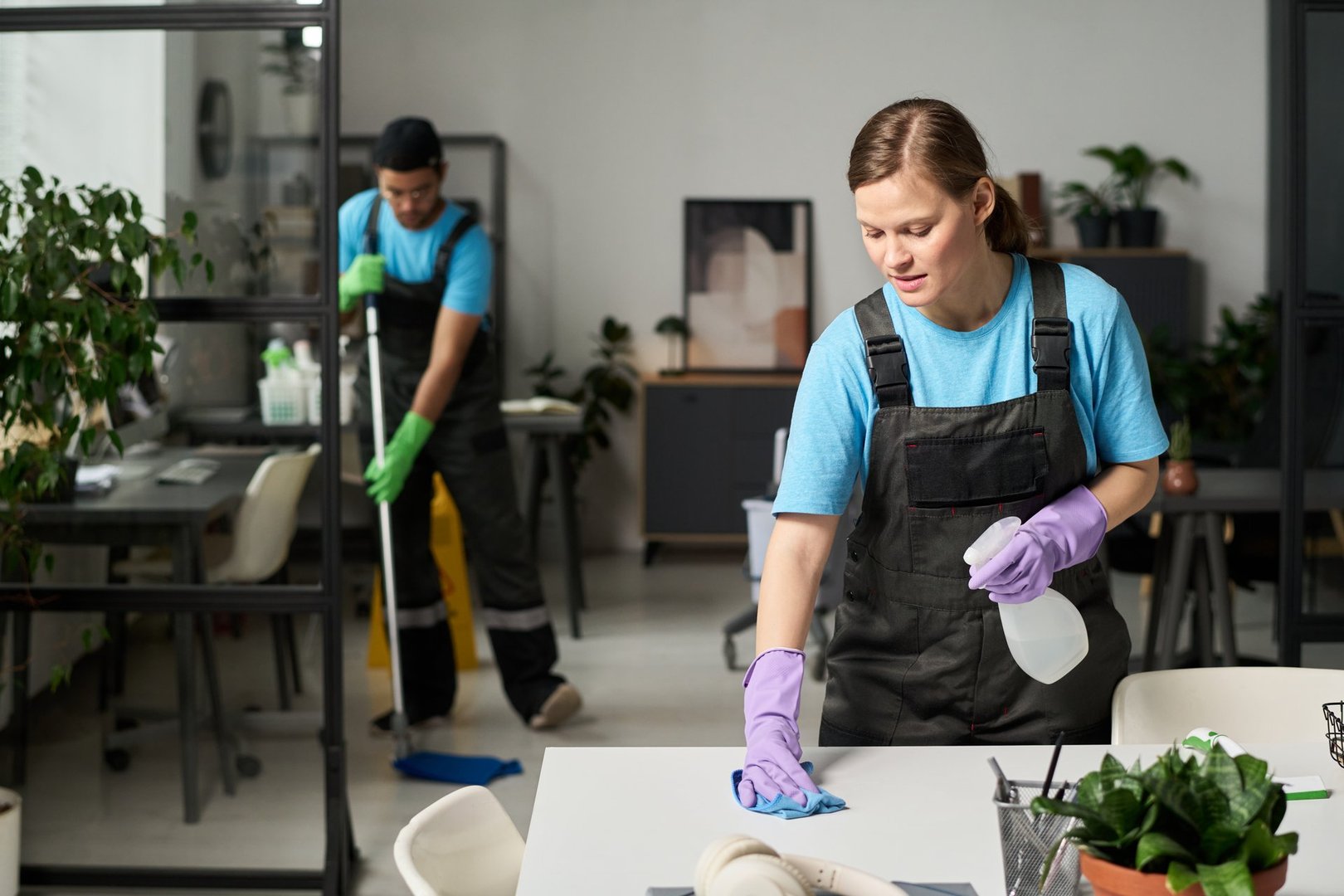 Woman in uniform wiping dust with rag and spray during her work with colleague in office
