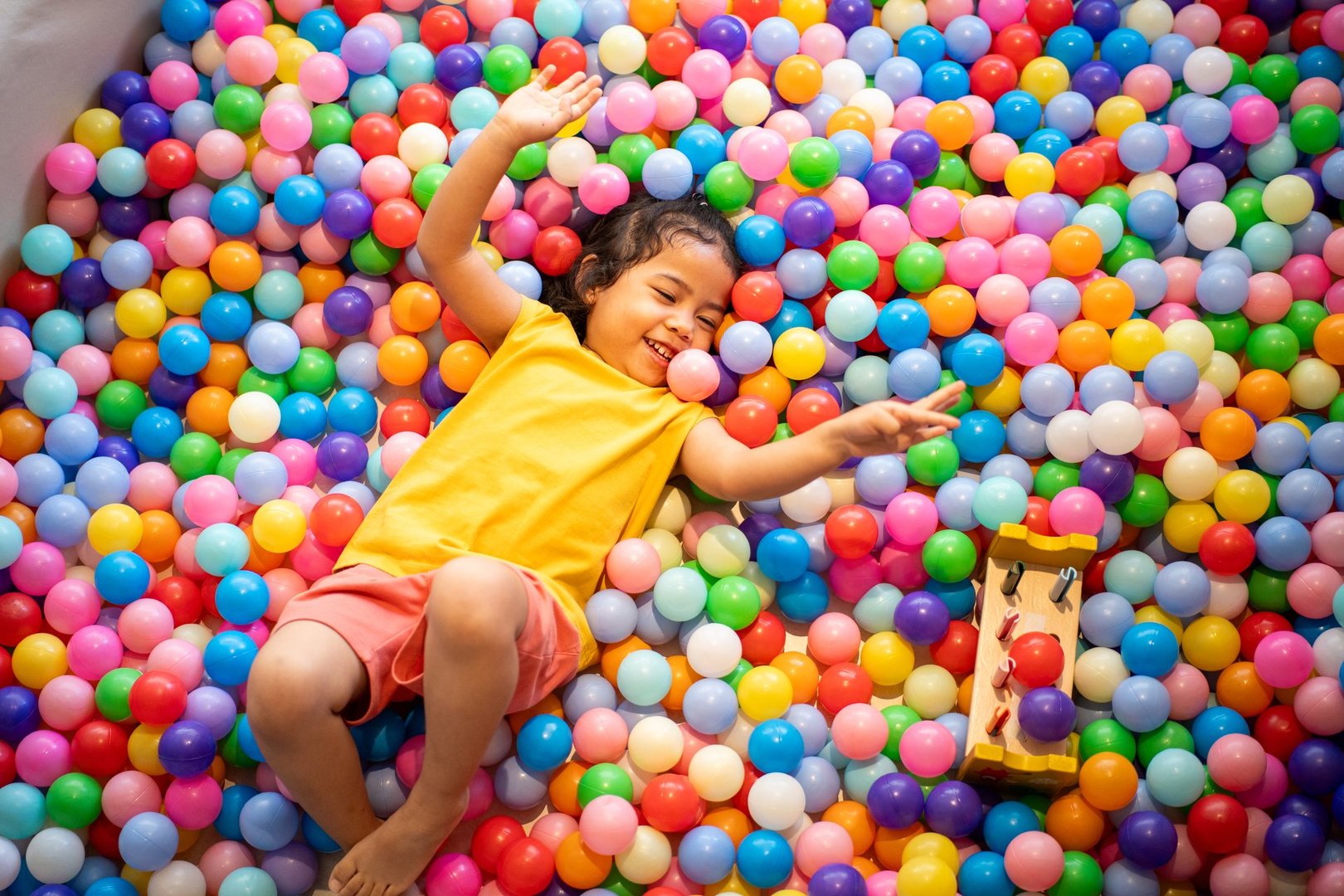 A happy little girl plays with a ball in a colorful ball pit, enjoying the fun and enhancing her development through playful activities and exercise.
