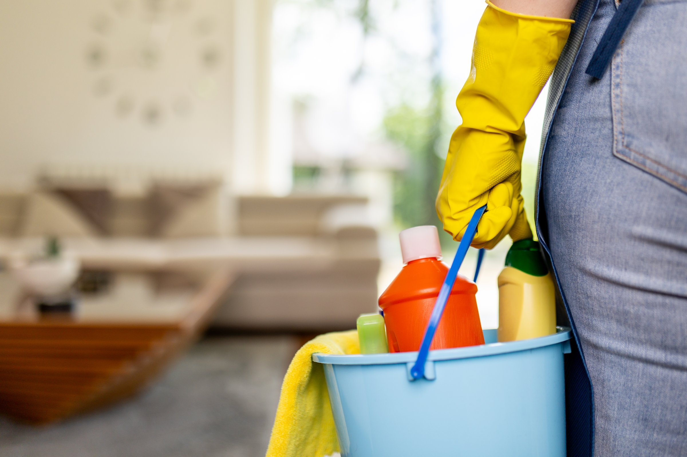 A person in yellow gloves holds a blue bucket filled with various cleaning supplies while preparing to start cleaning. Sunlight pours into the living area, highlighting the tidy environment.