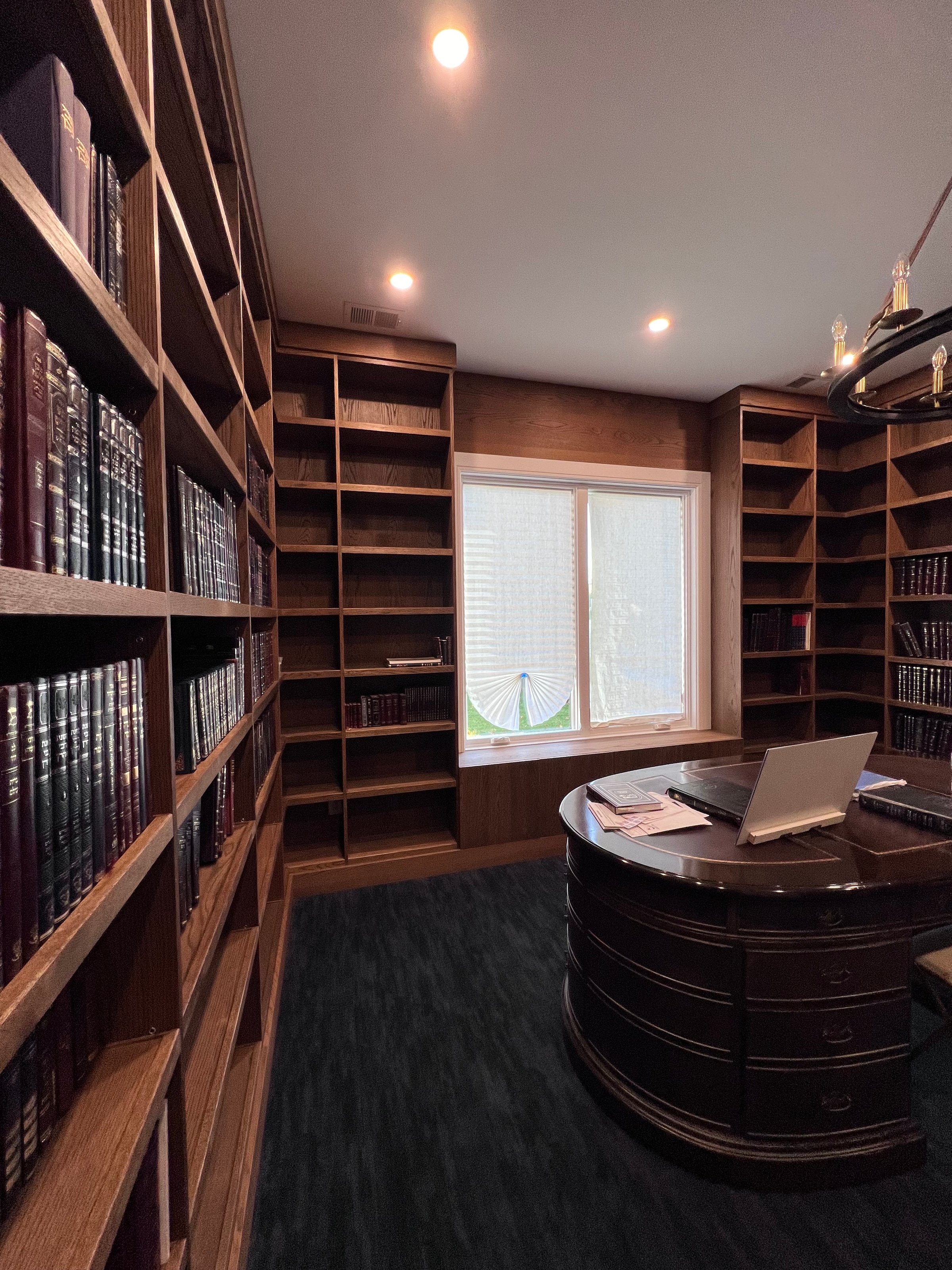 A cozy library room with wooden bookshelves, a large window, and a round desk with a laptop on it.