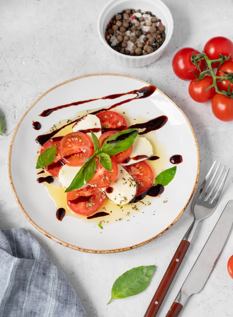 Fresh Caprese salad with sliced tomatoes, mozzarella cheese, basil leaves and balsamic glaze on a plate on a light background with fresh vegetables and spises. Healthy eating concept. Top view.