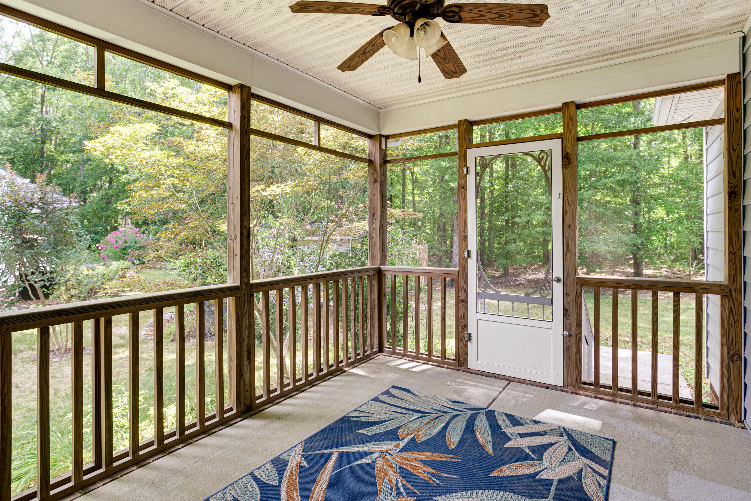A screened porch featuring a ceiling fan, wooden railing, and a lush green forest view in the background, creating a cozy and relaxing outdoor space.
