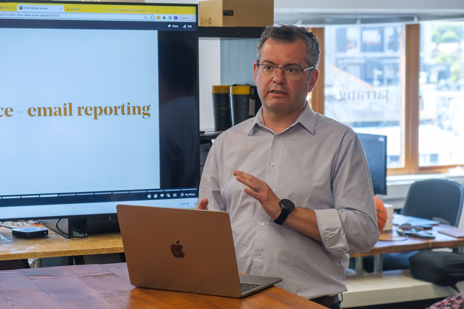 Person presenting in an office with a laptop and screen displaying email reporting. Casual attire and glasses.