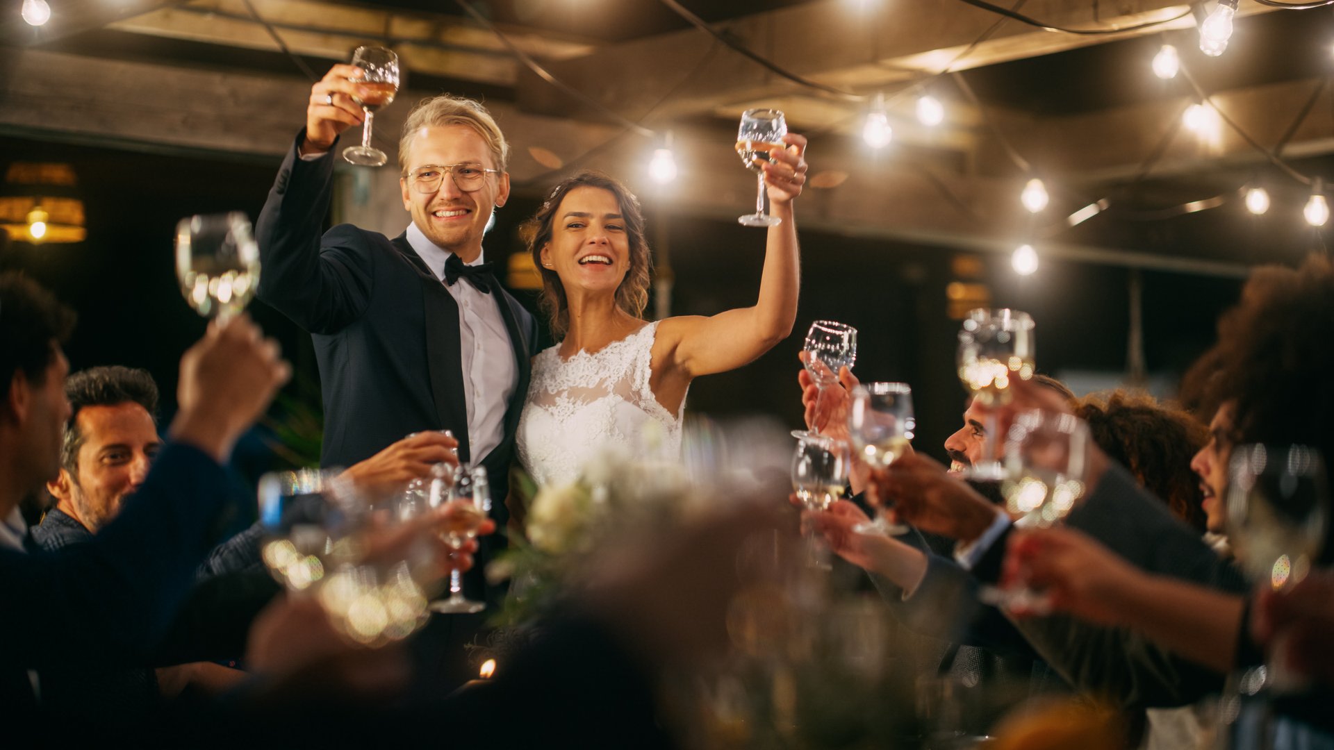 Beautiful Bride and Groom Celebrate Wedding at an Evening Reception Party. Newlyweds Propose a Toast to Happy Marriage, Standing at a Dinner Table with Best Multiethnic Diverse Friends.