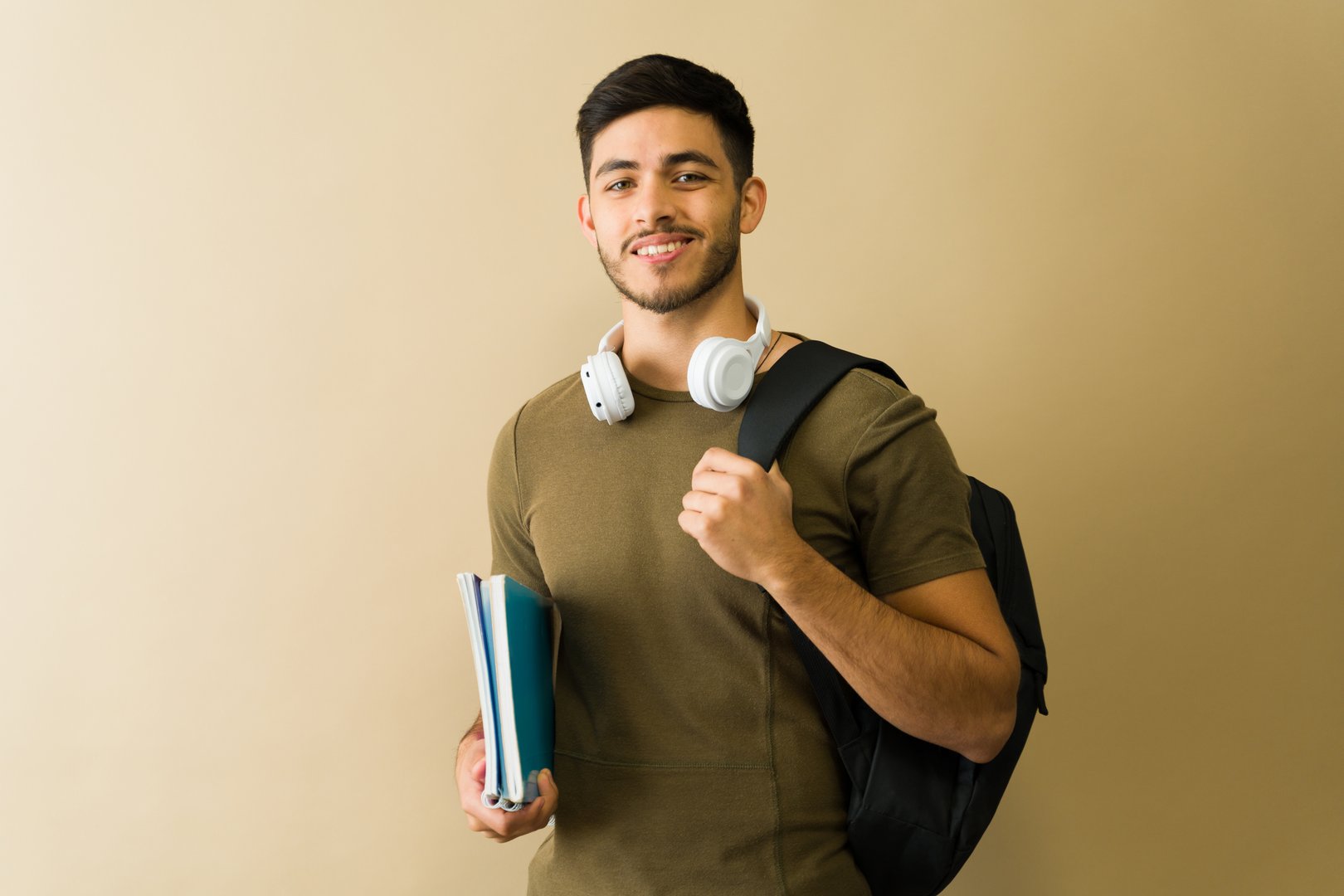Portrait of a smiling male university student carrying books and a backpack with headphones around his neck