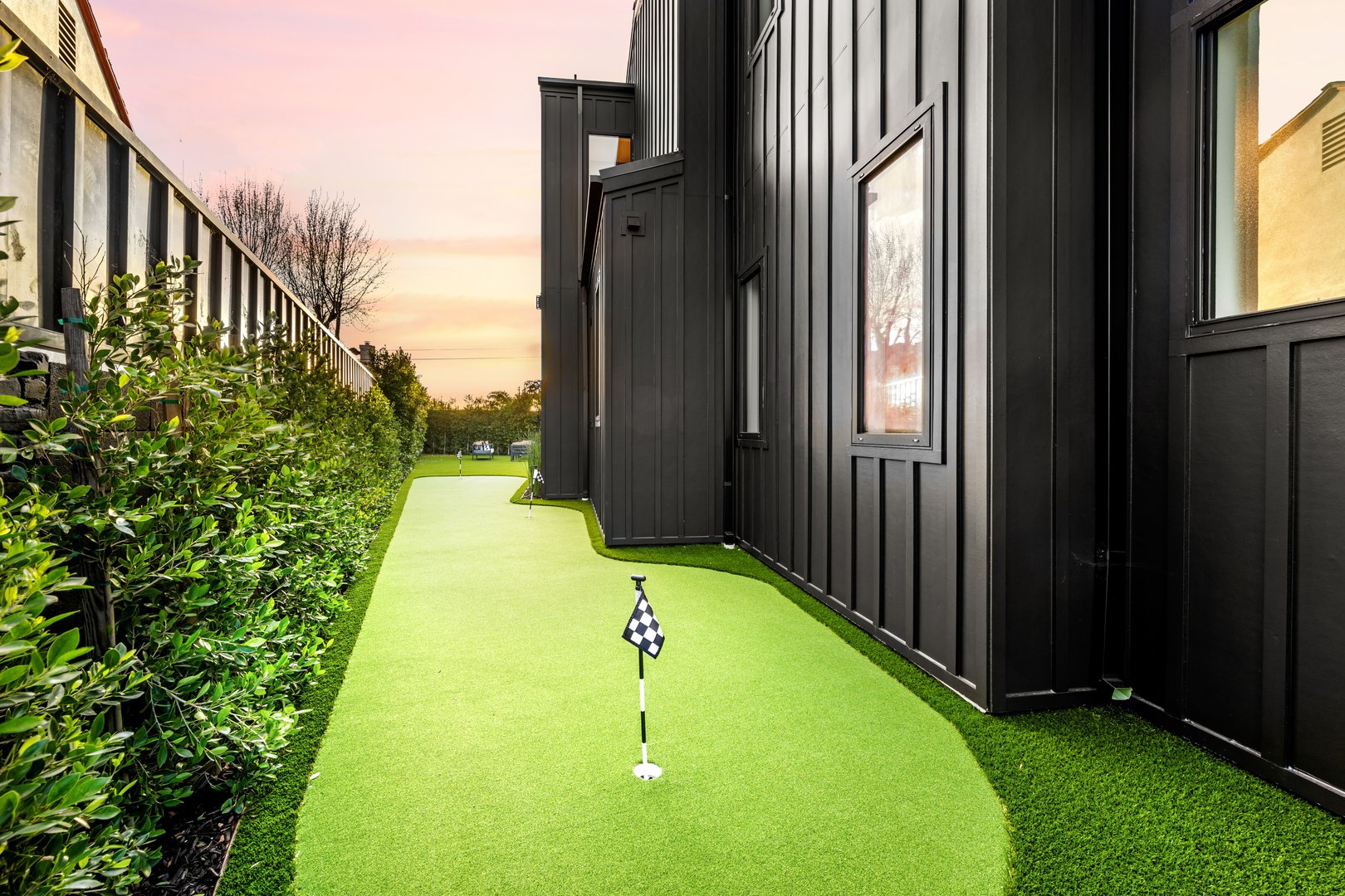 A backyard mini golf course with artificial turf next to a modern black house at sunset.