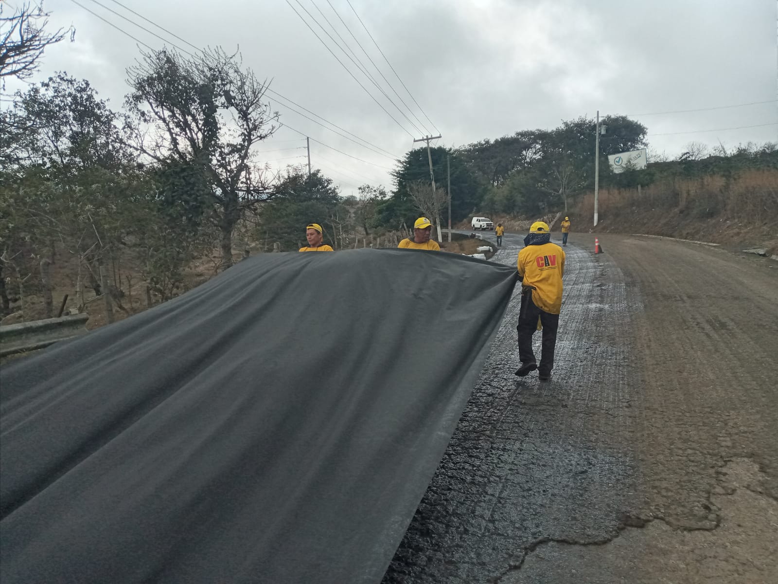 Workers in yellow uniforms lay down a large black sheet on a rural road under cloudy skies.