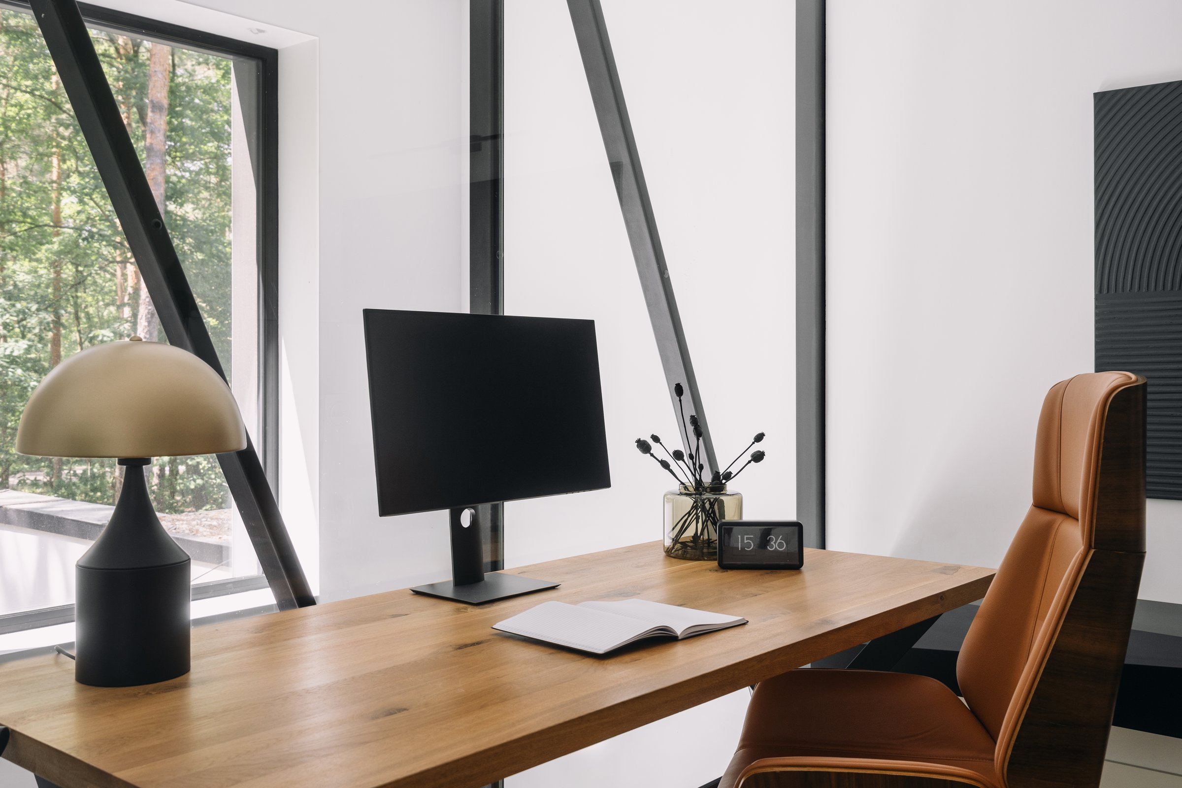 Contemporary workspace features a sleek wooden desk, computer monitor and elegant decor. Room with table lamp and a plant in space surrounded by large windows offering a view of nature in home office