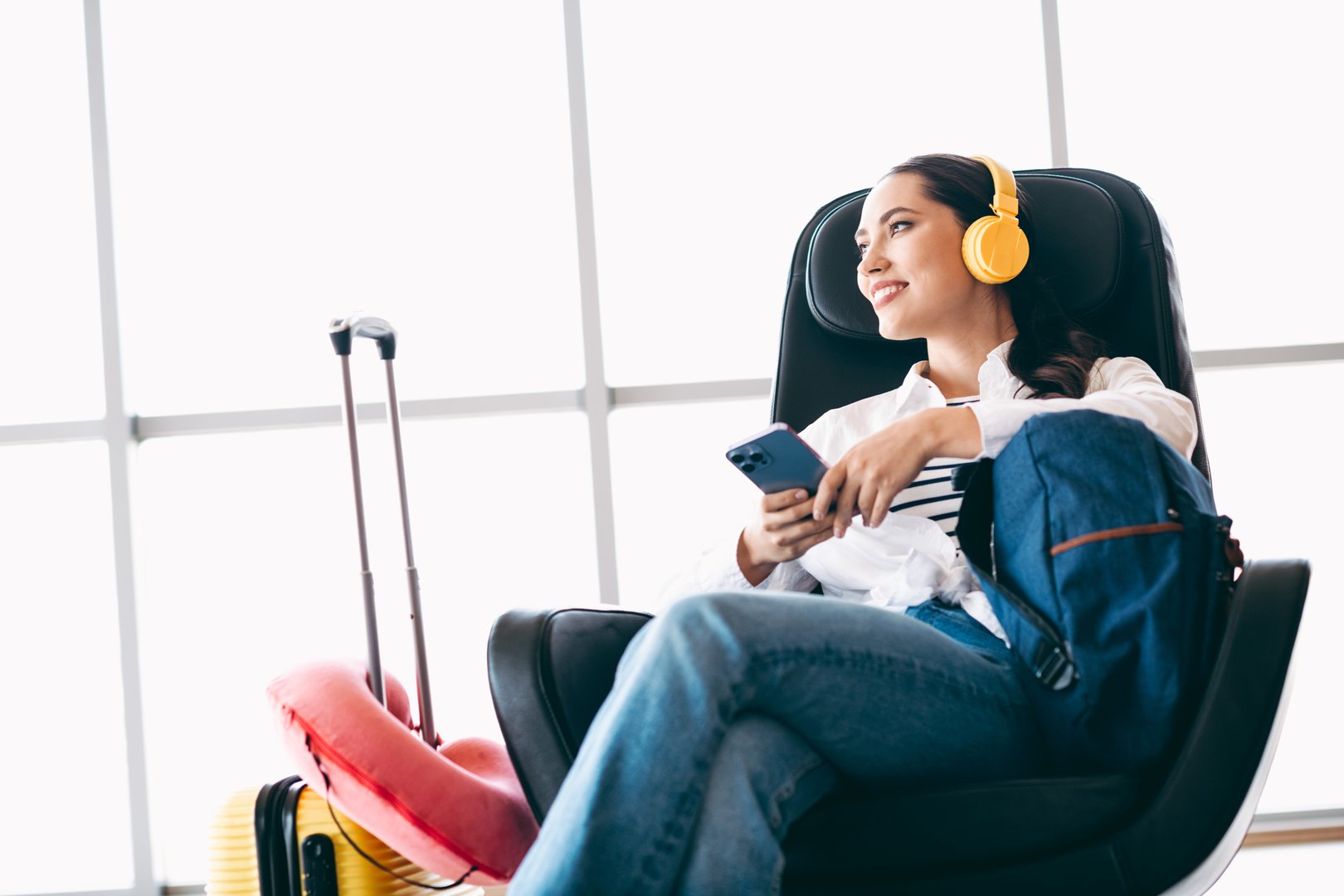 Female traveler at the airport wearing headphones, holding a phone, and seated with bags, anticipating her upcoming travel destination in a modern, casual setting.